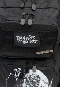 Čierny batoh má textúrovanú tkaninu, nastaviteľné popruhy a nášivky s nápismi "Číslo šelmy" a "Iron Maiden." Obsahuje grafický dizajn.