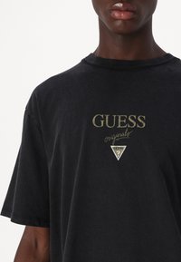 Svart bomull t-skjorte med korte ermer, med gullbrodert "GUESS originals" logo og grafikk av en trekant på brystet.