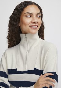 Pull crème avec des rayures marines, col zippé et texture côtelée. Présente un col montant et une coupe légèrement ample.