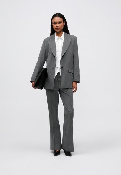 Un costume gris ajusté avec de larges revers, une chemise blanche en dessous et un pantalon à plis plats. Un sac à main noir est tenu dans la main gauche.
