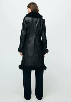 Abrigo de cuero negro con un cuello y puños de piel sintética, con un diseño ajustado y un acabado texturizado. Llevado sobre pantalones oscuros de pierna ancha.