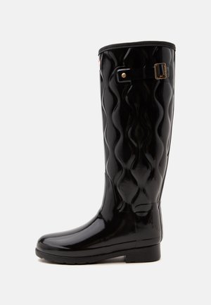 Bota de lluvia alta negra y brillante con un diseño texturizado ondulado y una pequeña hebilla lateral cerca de la parte superior.