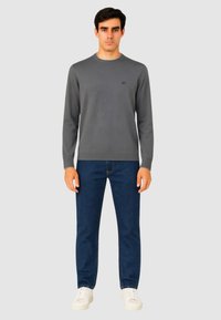Maglione grigio a maglia con scollo rotondo, abbinato a jeans blu scuro e sneakers bianche. Presenta un logo discreto sul petto.