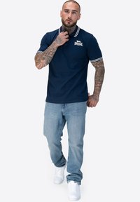 Marineblaues Poloshirt mit weißem gestreiften Kragen und Ärmeln, mit Logo auf der Brust. Kombiniert mit hellblauen Jeans und weißen Sneakern.