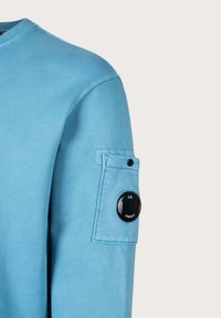 Camicia a maniche lunghe azzurro chiaro in cotone, con taschino sulla manica e un bottone nero rotondo con il marchio.