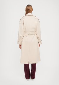 Beige Trenchcoat mit tailliertem Schnitt, großem Kragen und langen Ärmeln, aus glattem Stoff. Vertikaler Schlitz hinten für Bewegungsfreiheit.
