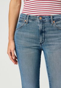 Jeans de mezclilla azul claro con cintura alta, diseño ajustado, bolsillos frontales, herrajes en tono cobre y un ligero desgastado en los muslos.