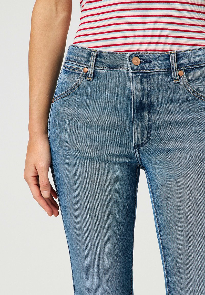 Jeans in denim leggermente blu con vita alta, design aderente, tasche frontali, accessori in tonalità rame e leggere sfumature sulle cosce.