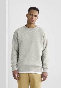 Grå hettegenser laget av fleece-materiale, med rund hals og ribbede mansjetter. Brukt over en hvit t-skjorte, med et merke på sømmen.