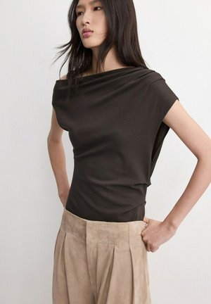 Femeie îmbrăcată într-un top fără mâneci, maro închis, cu un decolteu asimetric și pantaloni bej plisati, cu talie înaltă, stând cu mâinile pe șolduri.