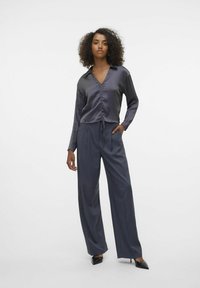 Grijze satijnen blouse met lange mouwen, een V-hals en een geplooide voorkant, gecombineerd met hoge grijze wijde broek en zwarte puntige hakken.