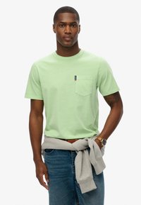 Lichtgroen katoenen t-shirt met een borstzak en een klein merklabel. Gecombineerd met blauwe jeans en een grijze sweatshirt om de taille geknoopt.