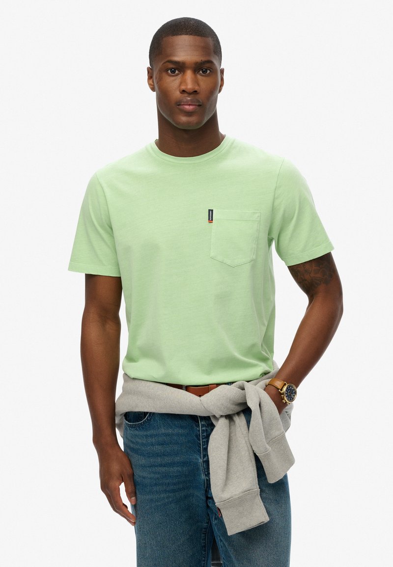 Lichtgroen katoenen t-shirt met een borstzak en een klein merklabel. Gecombineerd met blauwe jeans en een grijze sweatshirt om de taille geknoopt.