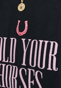 Černé tričko z látky s růžovým vyšitým textem "HOLD YOUR HORSES" a červeným grafickým prvkem podkovy, doplněné zlatým náhrdelníkem.
