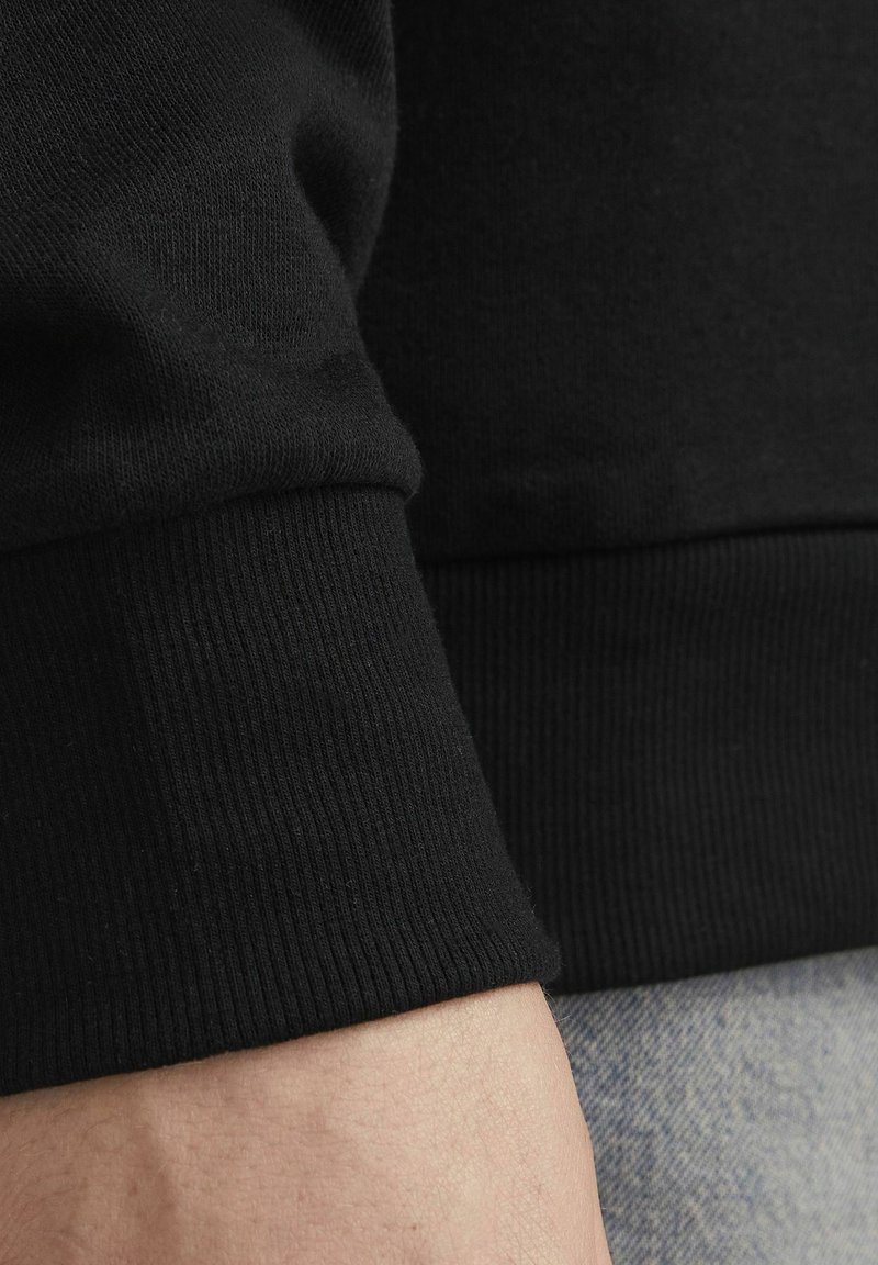 Polsino di felpa nera con texture a coste, in morbido cotone e con una finitura liscia, parzialmente visibile sopra un tessuto di denim.