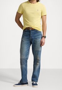 T-shirt di cotone gialla, jeans blu strappati con toppe, cintura in pelle marrone e sneaker in tela blu. Abbigliamento casual con dettagli minimi.