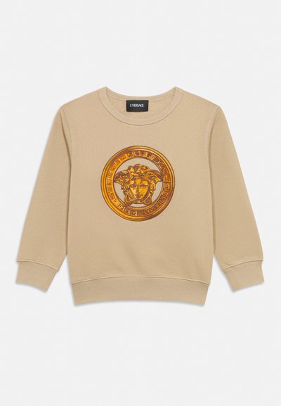 Sweat-shirt beige à manches longues avec un col rond, orné d'un grand emblème doré tête de Méduse dans une bordure circulaire décorative sur le devant.