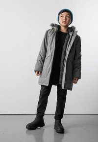 Chłopiec w szarej zimowej kurtce z futrzanym wykończeniem, czarnych spodniach, czarnych butach i niebieskiej wełnianej czapce, stojący na tle jednolitego tła.