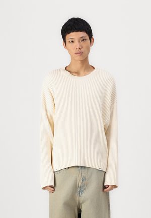 Pull crème côtelé à la coupe décontractée, col rond et épaules tombantes, associé à un pantalon en jean clair. Texture douce et design minimaliste.