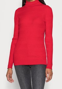Red ribbed turtleneck sweater with long sleeves, featuring a textured pattern and a fitted design. Pairing nicely with dark denim.