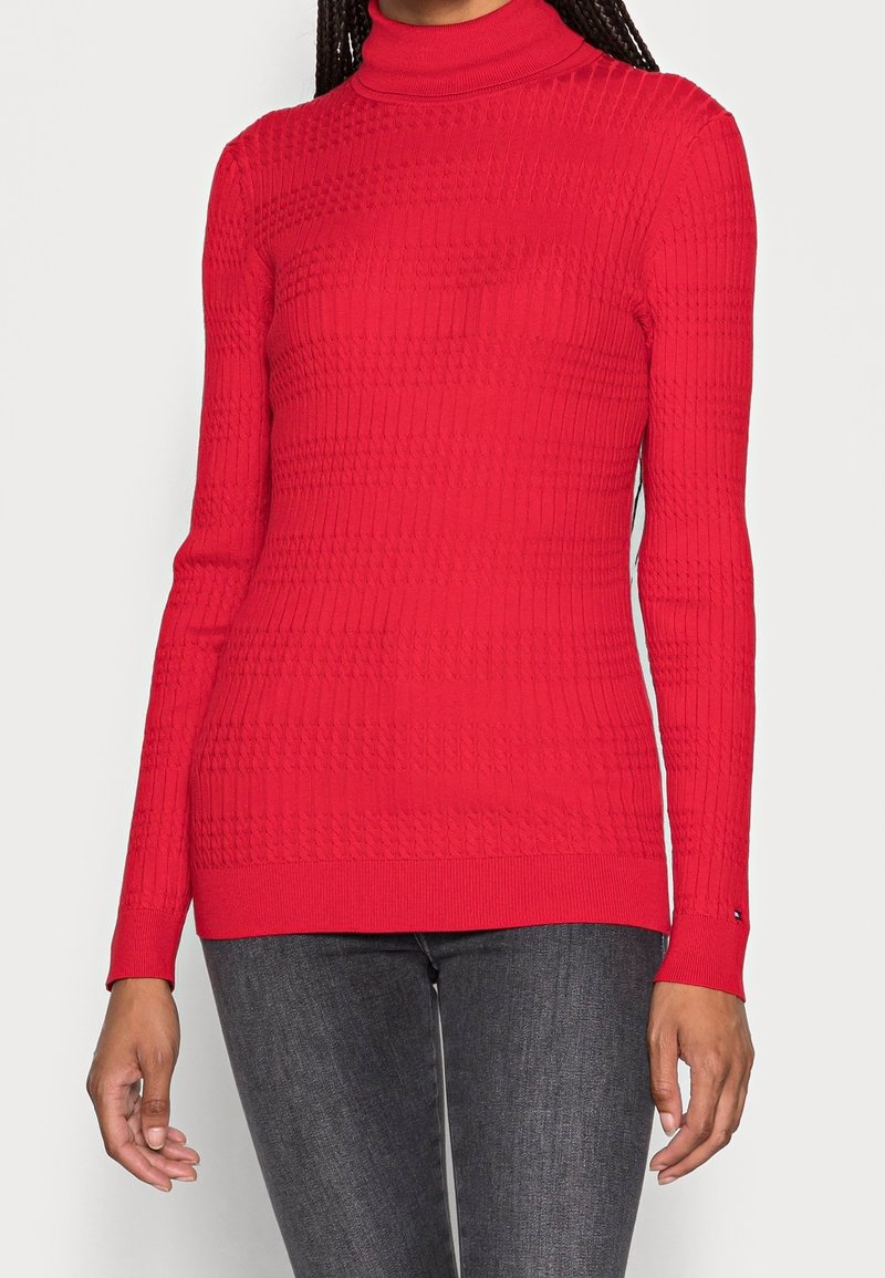 Red ribbed turtleneck sweater with long sleeves, featuring a textured pattern and a fitted design. Pairing nicely with dark denim.
