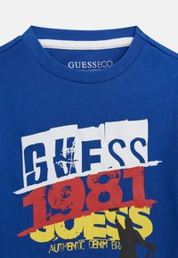 T-shirt in cotone blu con colletto a giro. Presenta una stampa grafica audace e multicolore con le scritte: "GUESS," "1981," e "AUTHENTIC DENIM."