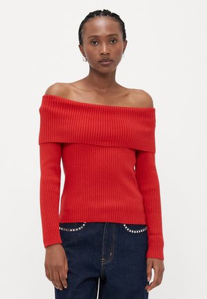Pull rouge à épaules dénudées en maille côtelée avec des manches longues et une coupe ajustée, associé à un jean foncé avec des accents décoratifs en argent.