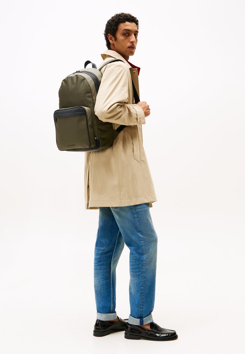 Grønn nylonryggsekk med en frontlomme, båret over en beige trenchcoat, kombinert med lys blå jeans og svarte blanke sko.