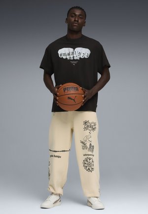 Mann trägt schwarzes Puma-T-Shirt und cremefarbene Hose mit schwarzen grafischen Mustern, hält einen braunen Puma-Basketball und steht vor grauem Hintergrund.