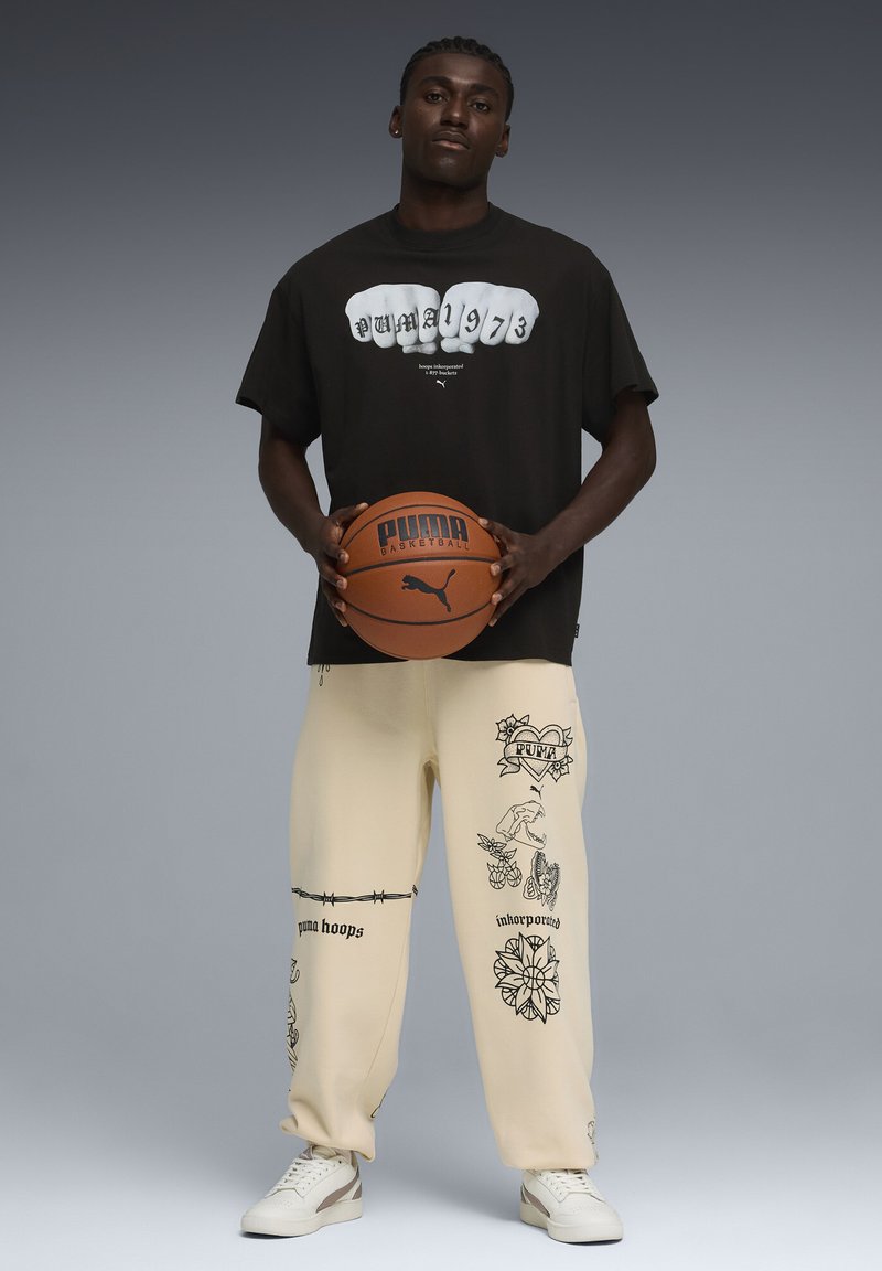 Άνδρας με μαύρο μπλουζάκι Puma και εκρού παντελόνι με μαύρα γραφικά σχέδια, κρατώντας καφέ μπάλα μπάσκετ Puma, στέκεται μπροστά σε γκρι φόντο.