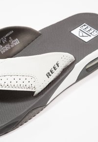 Tongs blanches et grises avec une tige perforée, semelle intérieure texturée et logo noir sur la lanière, fabriquées en matériaux synthétiques.
