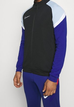 Man wearing a black zip-up jacket with light blue and white shoulders and blue sleeves paired with blue pants.
