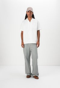 Camicia bianca a maniche corte con bottoni e tasca sul petto, abbinata a pantaloni a righe grigio e bianche e scarpe marroni. Cappellino beige chiaro sulla testa.