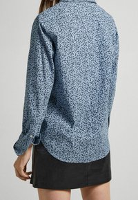 Camisa de botones azul con patrón, que presenta un dobladillo curvo, mangas largas y un tejido liviano con un diseño negro en contraste.
