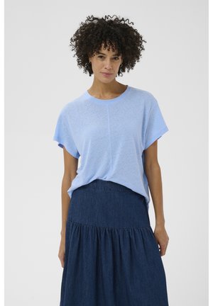 Femme aux cheveux bouclés portant un haut léger à manches courtes bleu clair, ample, rentré dans une jupe en denim bleu foncé taille haute, debout devant un fond uni.