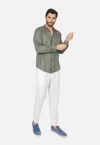 Camicia a maniche lunghe in lino verde con dettagli a bottoni, abbinata a pantaloni in lino bianchi e scarpe blu slip-on. L'uomo sta regolando la manica.