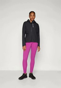 Donna in piedi con una felpa nera con zip, leggings viola brillante e scarpe da ginnastica nere su uno sfondo chiaro e neutro.