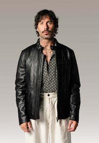 Veste en cuir noire avec un col montant et une fermeture éclair à l'avant, superposée à une chemise à motifs noir et blanc. Le mannequin a des tatouages visibles.