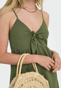 Robe verte olive avec un noeud torsadé sur le devant, fines bretelles et bas évasé. Le mannequin tient un sac rond en paille tissée avec deux poignées.