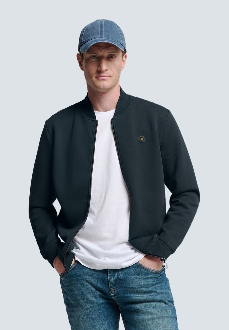 Dunkelgrüne Bomberjacke mit Reißverschluss, die mit einem sechseckigen Logo hervorsticht. Getragen über einem weißen T-Shirt und kombiniert mit blauen Jeans. Blauer Cap auf dem Kopf.