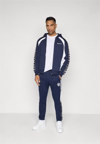 Fato de treino azul marinho com painéis laterais brancos e detalhes em faixas multicoloridas. Sweatshirt com fecho e logo no peito, combinada com calças de treino correspondentes.