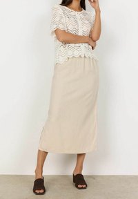 Žena ve bílém krajkovém topu s krátkým rukávem a knoflíky, v kombinaci s béžovou midi sukní a černými sandály na podpatku s odkrytými prsty.