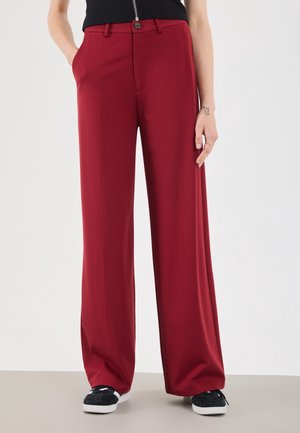 Persona que lleva pantalones rojos de tiro alto y pierna ancha con bolsillos, combinados con zapatillas negras y blancas y un top negro con cremallera frontal.