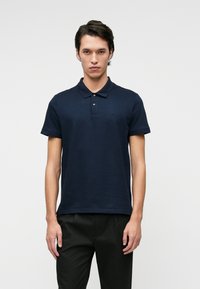 Tengerészkék pamut póló ing három gombos gallérral, rövid ujjakkal és diszkrét logóval a mellkasán. Fekete nadrággal viselve.