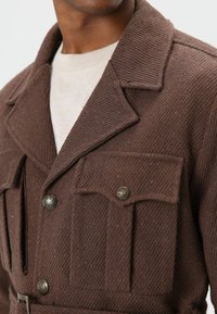 Braune Jacke mit Struktur, mit Kragen, zwei Brusttaschen und metallischen Knöpfen, getragen über einem hellfarbenen Pullover.