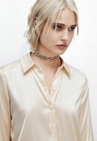 Camicia in satin color crema con patta dei bottoni e colletto a punta. Indossata con un collare choker in argento con dettagli geometrici.