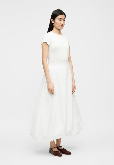 Γυναίκα με μακριά μαύρα μαλλιά φοράει λευκό τοπ με κοντά μανίκια και φουσκωτή λευκή midi φούστα, με καφέ παπούτσια Mary Jane, στέκεται πλαγίως.