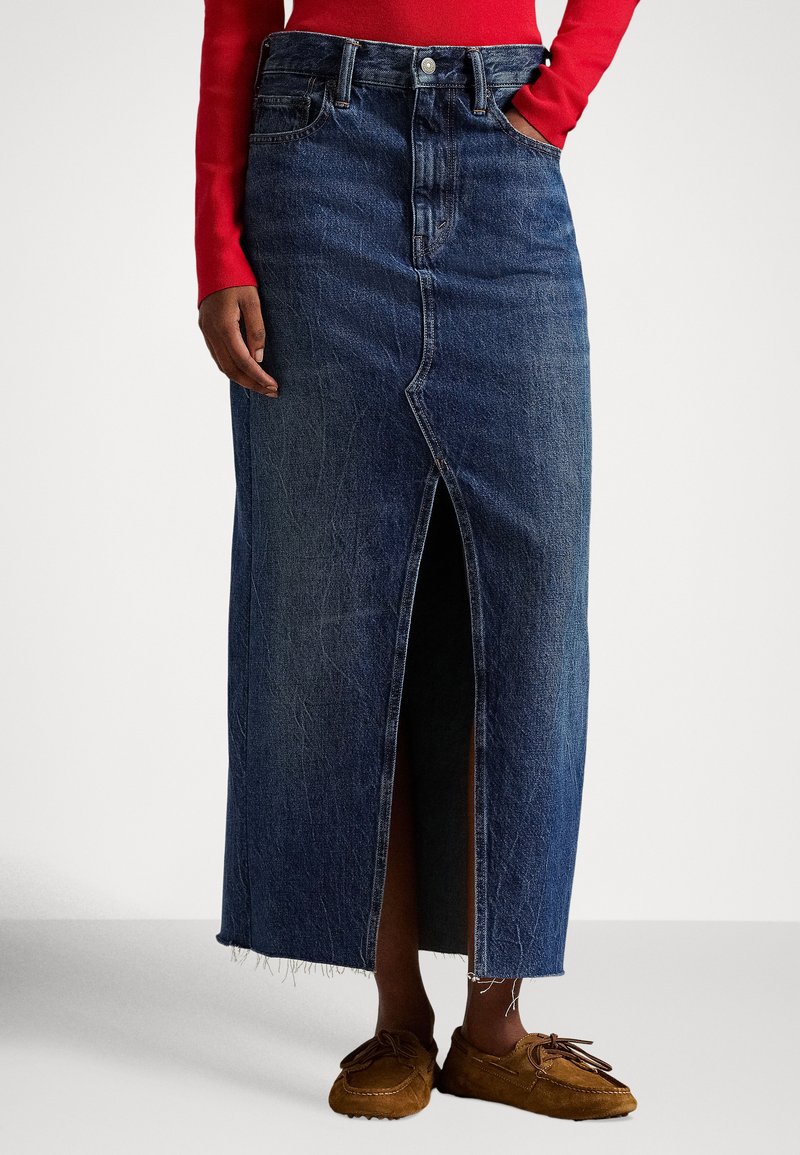 Osoba stojaca pred jednoduchým pozadím, oblečená v dlhej modrej rifľovej sukni s predným rozparkom, červenom tričku s dlhým rukávom a hnedom mokasínov.