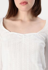 Haut blanc à manches longues avec un décolleté carré. Présente un motif texturé avec des rayures verticales et de petits détails en œillets. Légère fronce au centre.