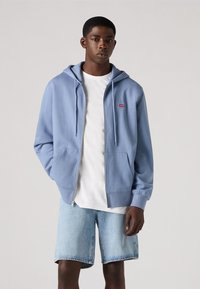 Lichtblauwe zip-up hoodie met een grote voorkantzak, trekkoordcapuchon en rode logopatch. Draagt over een wit T-shirt en blauwe denimshorts.